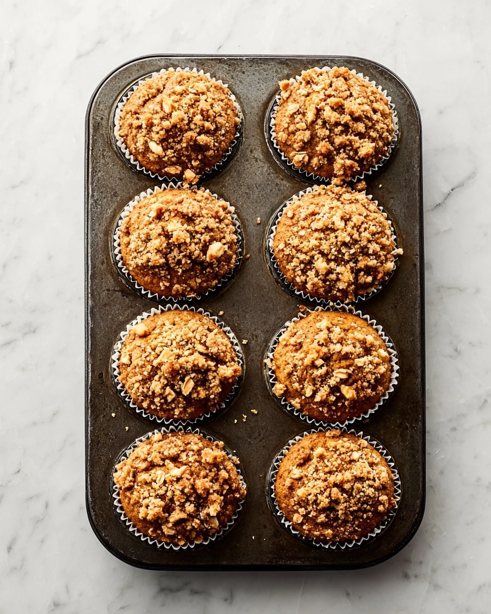 A metal muffin tray holds eight freshly baked muffins, each topped with a crumbly, golden-brown oat streusel that adds a rough texture on top. The muffins themselves are a light brown color with a slightly domed shape, visible through white paper liners. The tray rests on a white marbled surface that contrasts gently with the warm tones of the muffins and the dark metal pan. The muffin tops have small clusters of oats and crumbs scattered unevenly, giving a rustic homemade look. Photo taken with an iphone --ar 4:5 --v 7