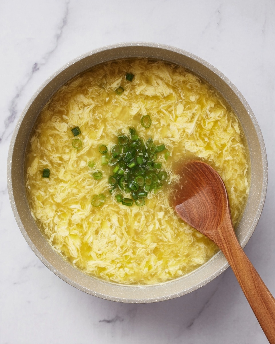 A round light gray pot filled with yellow egg drop soup shows a textured surface with soft, wavy egg strands floating in clear broth. In the center, there is a small pile of finely chopped bright green scallions adding a fresh touch. A wooden spoon with a smooth light brown handle rests inside the soup on the right side. The pot sits on a white marbled surface, enhancing the colors of the soup. photo taken with an iphone --ar 4:5 --v 7