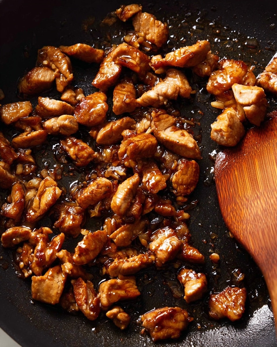 This image shows small pieces of cooked meat in a dark non-stick pan. The meat is golden brown with a shiny surface, showing that it is cooked with some oil or sauce. A wooden spatula is on the right side of the pan, partially under some meat pieces, with a smooth texture and some oil on it. The pan’s background is dark and slightly glossy from the oil. The image is close up, focusing on the cooked meat texture and the spatula. Photo taken with an iphone --ar 4:5 --v 7