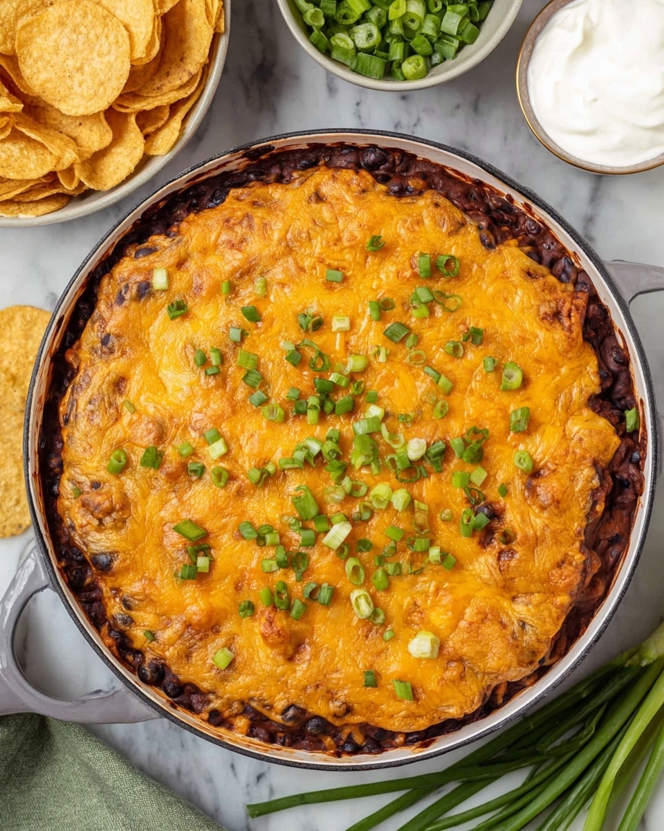 A round white pan filled with a layered dish starting with a dark, thick base layer that looks like black beans, topped by a thick, golden brown cheesy layer that covers the whole surface, sprinkled with small pieces of chopped green onions spread evenly on top. Around the pan are a bowl of round, golden-brown chips and a small white bowl with smooth white sauce, all placed on a white marbled surface with some chopped green onions in the background. photo taken with an iphone --ar 4:5 --v 7