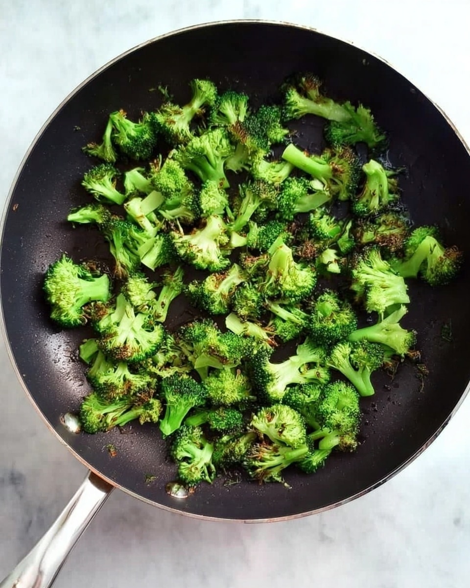 A black frying pan filled with small pieces of bright green broccoli that are slightly cooked, showing some darker, lightly browned edges. The broccoli pieces are spread out evenly in a single layer inside the pan. The handle of the pan is silver and visible at the bottom left of the image, resting on a white marbled surface. Photo taken with an iphone --ar 4:5 --v 7