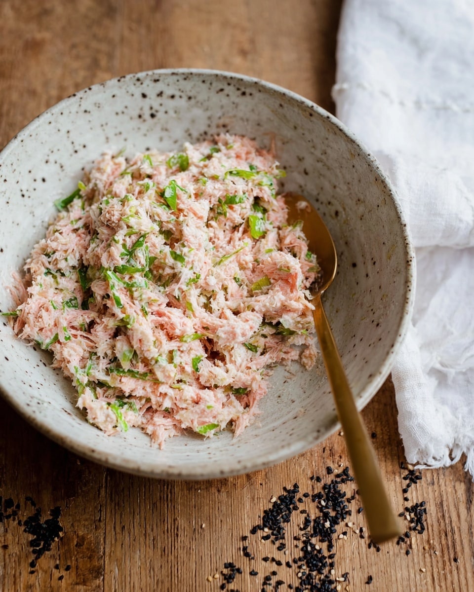 A white speckled bowl holds a shredded mixture with a light pink color, mixed with small pieces of green herbs, giving it a fresh look. The texture appears soft and finely shredded, with visible bits of green evenly spread throughout. A gold spoon rests inside the bowl on the right side, slightly buried in the mixture. The bowl is placed on a wooden surface with some black seeds scattered around, and a white cloth is partly visible in the background. photo taken with an iphone --ar 4:5 --v 7