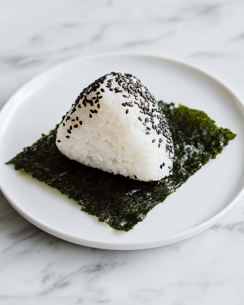 A single triangular-shaped rice ball is placed on top of a dark green, textured seaweed sheet that lays flat on a white plate, which is set against a white marbled surface. The rice ball is white with black sesame seeds sprinkled evenly on its outer surface, giving it a speckled appearance. The seaweed sheet underneath the rice ball has a slightly shiny and rough texture, contrasting with the smooth, soft look of the rice layers. Photo taken with an iphone --ar 4:5 --v 7