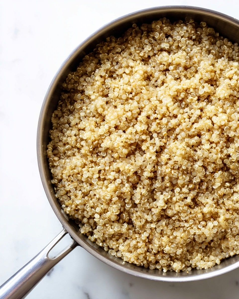 A close-up view of a silver metal pan filled with cooked quinoa grains. The quinoa is light beige to golden color with a soft, slightly fluffy texture. The grains fill the pan evenly, showing their round shape and tiny white curls. The pan is placed on a white marbled surface, and its handle extends outwards towards the bottom left of the image. The image is bright with clear details of the quinoa. photo taken with an iphone --ar 4:5 --v 7