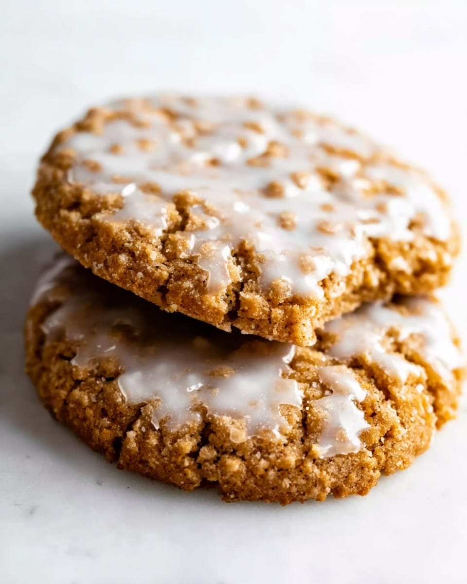 The image shows two round oatmeal cookies stacked on a white marbled surface. The bottom cookie is fully visible with a golden-brown texture and rough, crumbly edges. The top cookie is placed diagonally on the bottom one and has a similar texture. Both cookies are covered with thin, white icing that spreads unevenly across the top, creating a light glistening effect with some small cracks and bubbles. The overall look is rustic and homemade. photo taken with an iphone --ar 4:5 --v 7