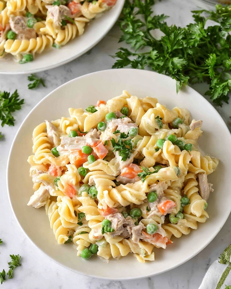 The image shows a close-up of a white plate filled with creamy rotini pasta salad. The pasta is twisted and light yellow, mixed with small, bright green peas and small orange carrot cubes. There are also pieces of light beige cooked meat scattered throughout. The salad looks creamy and well mixed with a few small pieces of green parsley sprinkled on top. The plate sits on a white marbled surface with some green parsley leaves nearby. Another similar plate partly appears in the background. photo taken with an iphone --ar 4:5 --v 7