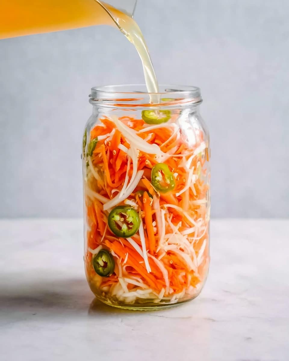 The image shows a clear glass jar filled with thin, long strips of white and orange vegetables, mostly layered in an even mix. Small slices of green chili peppers are evenly placed around the inside of the jar, visible against the colorful strips. A liquid, pale yellow in color, is being poured into the jar from the top left corner. The jar is placed on a white marbled surface, and the background is a soft light color. The scene looks clean and fresh, with a focus on the bright colors and textures inside the jar. photo taken with an iphone --ar 4:5 --v 7