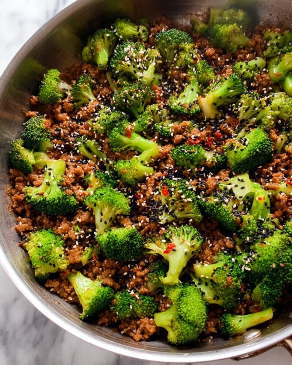 A close-up view of a silver pan filled with a dish that has two main layers: bright green broccoli florets with a slightly charred texture scattered evenly on top, and a layer of finely chopped cooked brown meat sauce beneath, mixed throughout with small bits of red pepper flakes and garnished with black and white sesame seeds sprinkled across the entire surface. The pan is set on a white marbled background. photo taken with an iphone --ar 4:5 --v 7