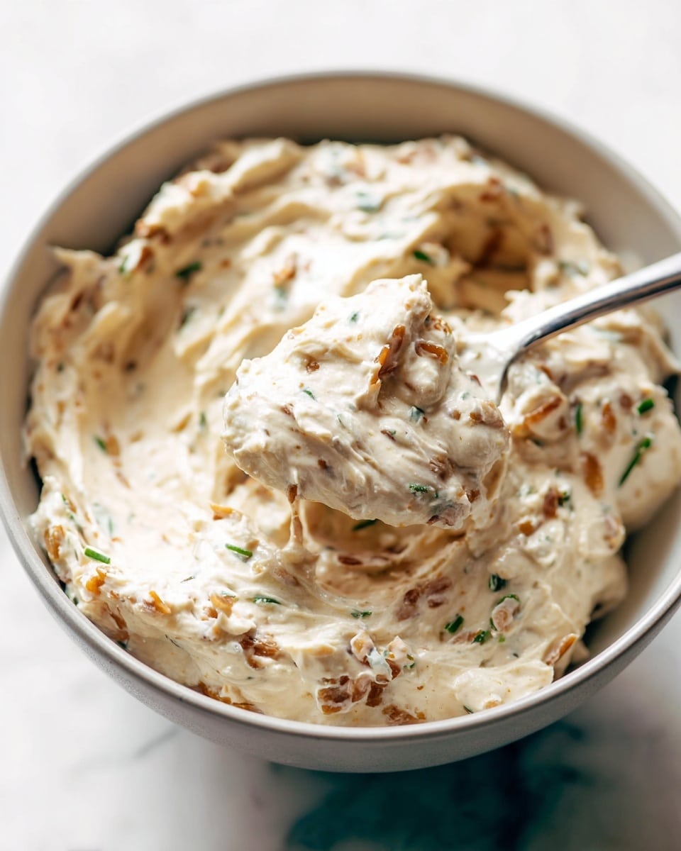 A close-up view of a creamy dip inside a white bowl, showing soft, thick texture with visible small pieces of herbs and finely chopped light brown ingredients mixed through it. A silver spoon is scooping some of the dip, emphasizing its smooth yet chunky consistency. The bowl rests on a white marbled surface, and the parfait’s colors range from off-white to light brown with hints of green from the herbs. photo taken with an iphone --ar 4:5 --v 7