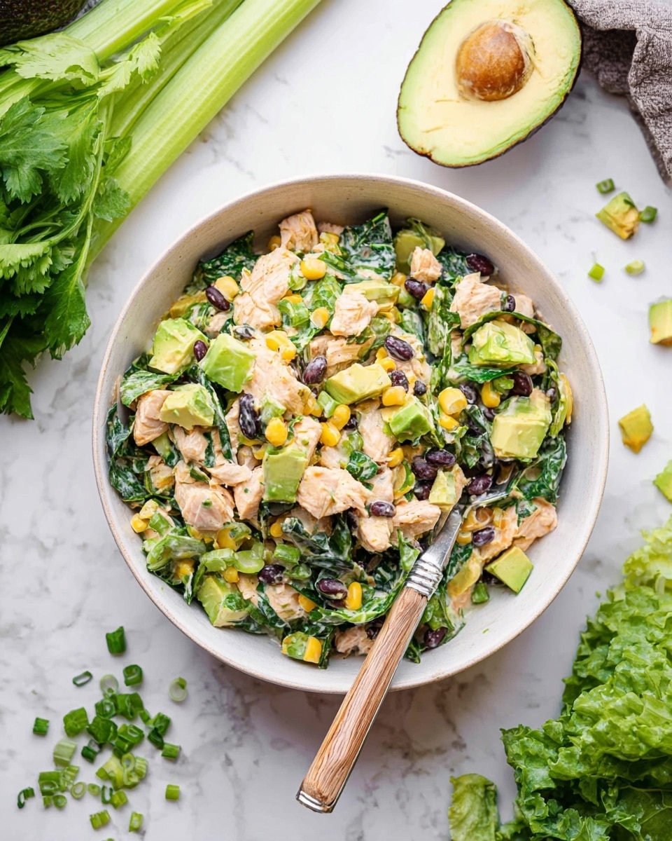 The image shows a white bowl filled with a fresh salad that has about four main layers: chunks of light pink cooked chicken mixed with diced light green avocado, shiny black beans, bright yellow corn, and chopped dark green leafy vegetables, all tossed together in a creamy dressing. The bowl sits on a white marbled surface with a silver fork with a wooden handle resting inside the bowl, partially buried in the salad. Surrounding the bowl are fresh ingredients like half an avocado with its brown seed, green celery stalks, dark leafy greens, and some scattered small pieces of yellow bell pepper and chopped green onions. Photo taken with an iphone --ar 4:5 --v 7
