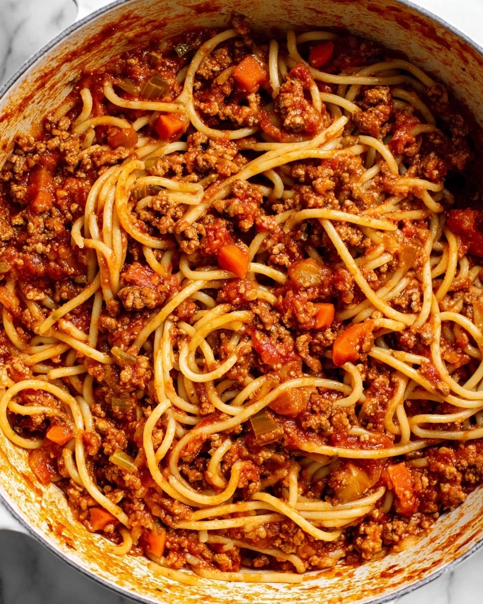 The image shows a close-up view of a pot filled with spaghetti mixed with ground meat sauce. The spaghetti noodles are light brown and soft, tangled throughout the pot. The sauce is a rich red-orange color with chunks of cooked ground meat and small pieces of orange carrots and green celery scattered evenly. The pot is white, and traces of sauce stick to its sides. The background surface is a white marbled texture. Photo taken with an iphone --ar 4:5 --v 7
