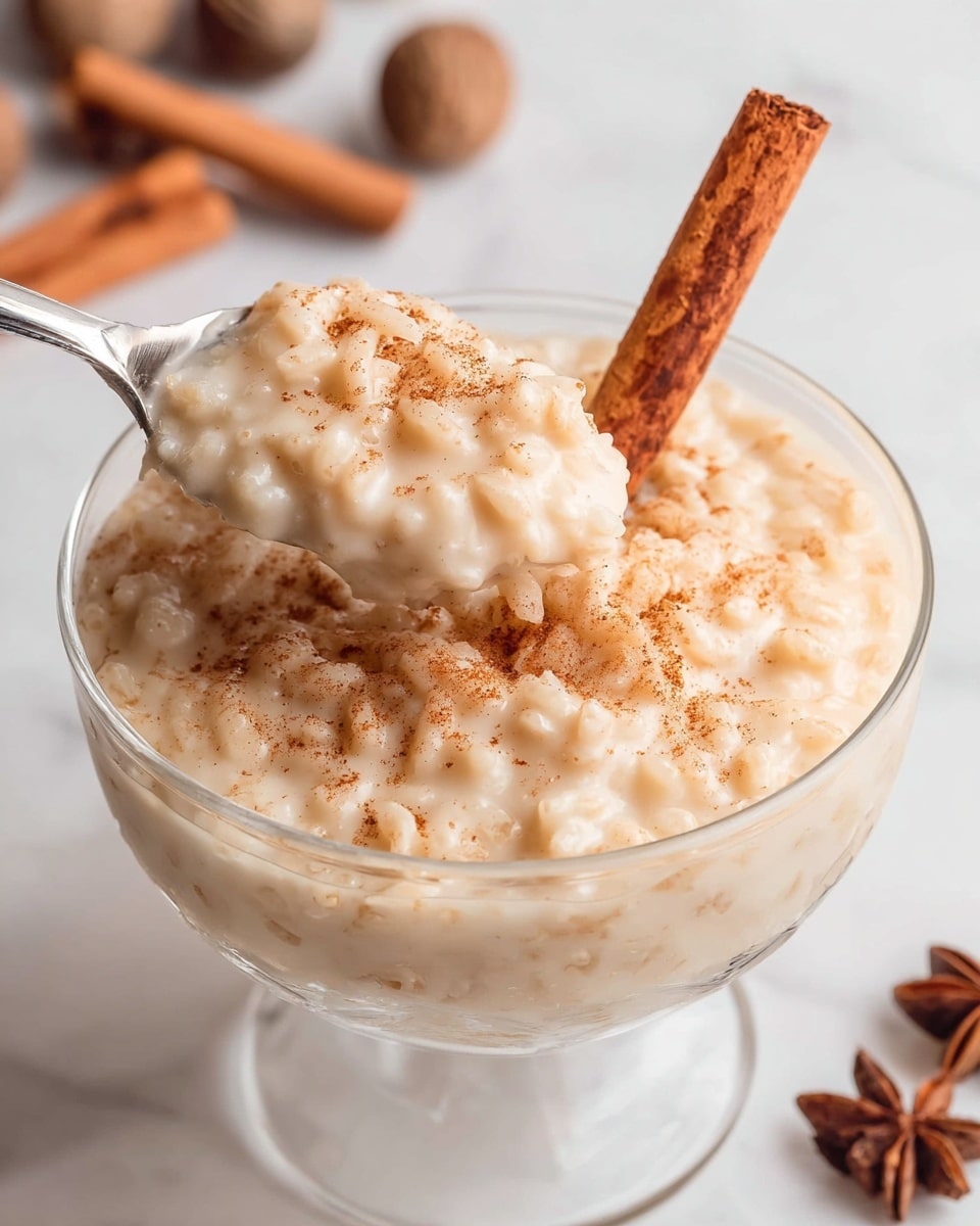 A clear glass bowl filled with thick, creamy rice pudding that has a pale beige color. The pudding is textured with visible small rice grains, slightly piled above the rim of the bowl. A silver spoon is scooping some pudding from the bowl, showing the creamy, slightly lumpy texture. The top of the pudding is sprinkled lightly with brown cinnamon powder. A cinnamon stick is placed upright inside the pudding towards the back. The bowl sits on a white marbled surface with blurred cinnamon sticks and whole nutmegs in the background. Photo taken with an iphone --ar 4:5 --v 7
