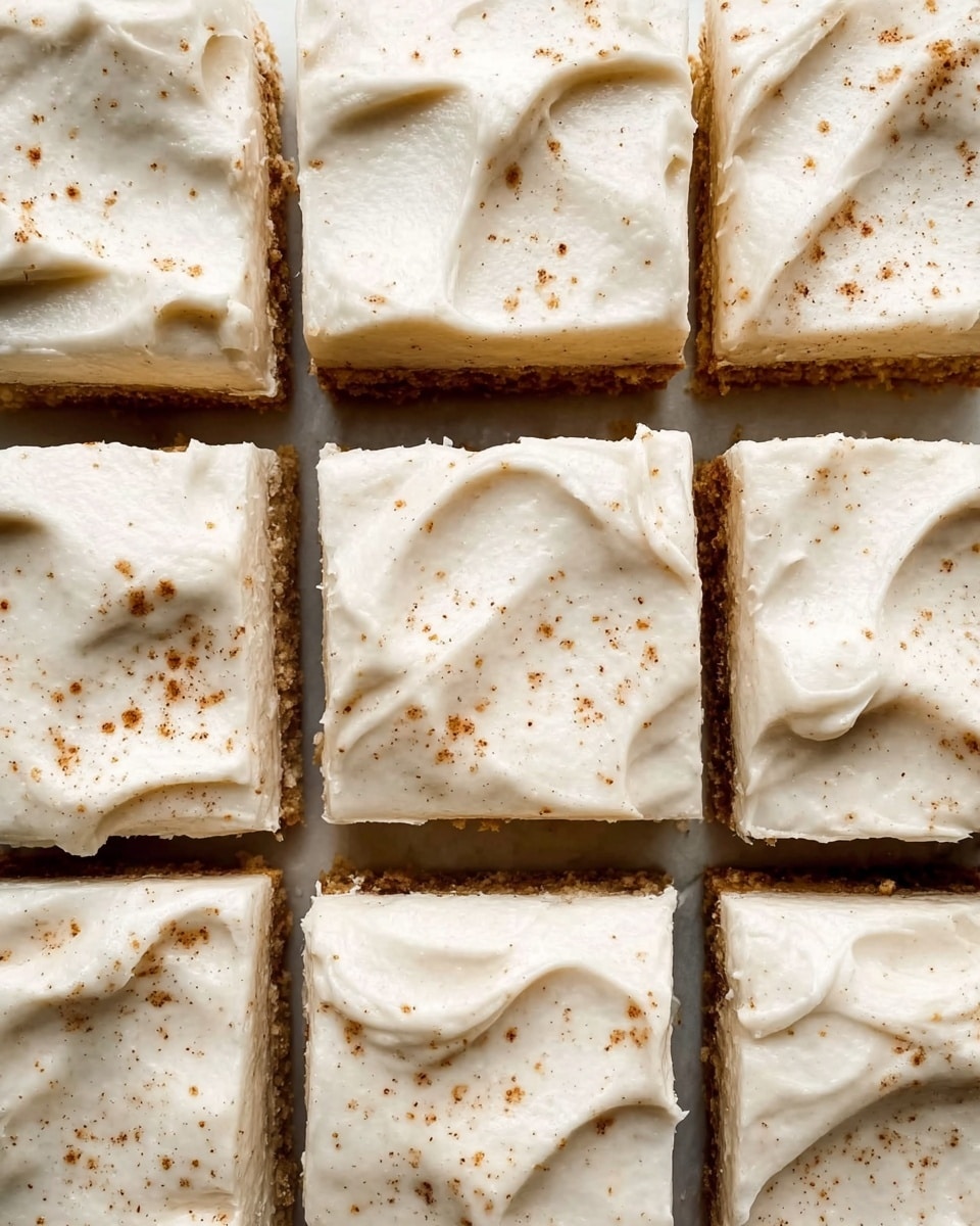 The image shows a close-up view of a batch of square-shaped, frosted cake pieces arranged in a grid. Each piece has a smooth, creamy white frosting layer with slight swirls and small specks of light brown sprinkled on top, likely cinnamon or nutmeg. Beneath the frosting, glimpses of a darker cake layer can be seen where the frosting is thinner or has cracks. The cake pieces are placed against a white marbled background, with soft natural light highlighting the texture of the frosting. photo taken with an iphone --ar 4:5 --v 7