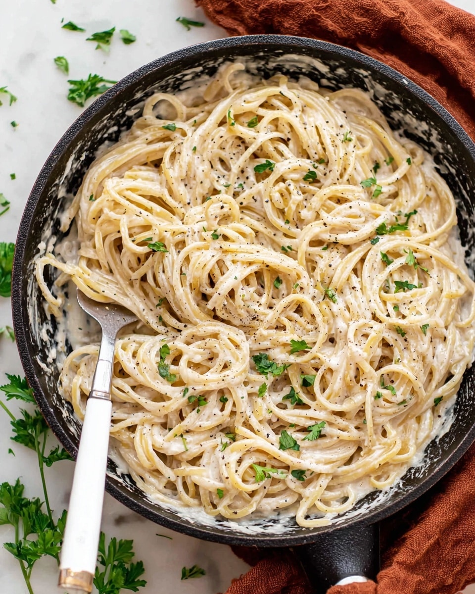 A black skillet filled with spaghetti covered in a creamy white sauce with small bits of black pepper visible throughout. The pasta strands are thick and intertwined, coated evenly with the sauce. Fresh green parsley leaves are sprinkled on top, adding a touch of color. A fork with a white handle rests inside the skillet, twirling some spaghetti. The skillet is placed on a white marbled surface, with some parsley leaves scattered nearby and part of a rust-colored cloth peeking out from under the skillet. Photo taken with an iphone --ar 4:5 --v 7