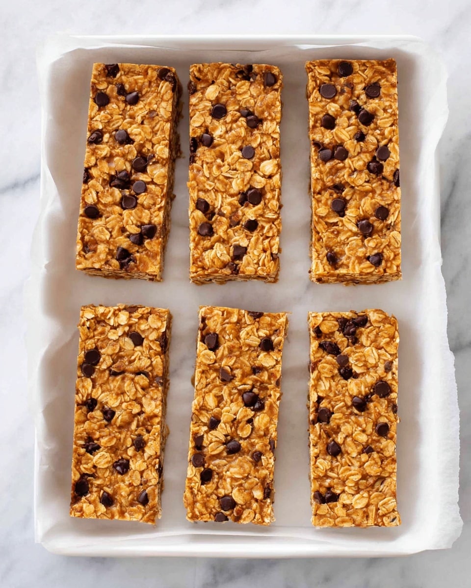 Six rectangular granola bars are placed on white parchment paper inside a white tray. Each bar is thick and golden brown with a rough texture showing oats and small dark chocolate chips evenly spread throughout. The edges are clean and straight, and the bars are arranged in two rows of three on a white marbled surface. There is no other decoration or extra items in the scene, keeping the focus on the oat and chocolate mixture in the bars. photo taken with an iphone --ar 4:5 --v 7
