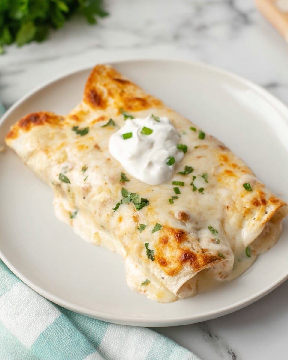 A single folded enchilada sits on a white plate, covered in creamy, lightly browned cheese sauce with small green herb bits scattered on top. The enchilada shows a soft, smooth texture with some slight bubbling of the cheese. On top is a dollop of white sour cream, garnished with small green herb pieces. The background is a white marbled surface with a pale blue and white checkered cloth partially visible at the bottom edge. Photo taken with an iphone --ar 4:5 --v 7