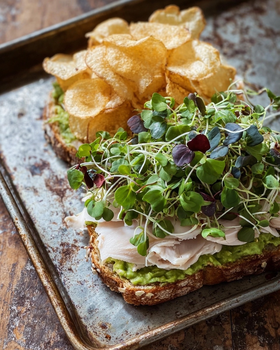 The image shows an open sandwich on toasted brown bread placed on a rustic metal tray over a wooden surface. The bottom slice has green smashed avocado spread as the first layer, topped by thin slices of pale white turkey arranged in soft folds. On top of the turkey, there is a pile of light golden, slightly crispy potato chips. The top slice of bread is covered with fresh, bright green microgreens with a few dark purple leaves mixed in, giving a fresh and textured look. The sandwich layers show a mix of soft, creamy, and crunchy textures with earthy colors, all seen from above. photo taken with an iphone --ar 4:5 --v 7