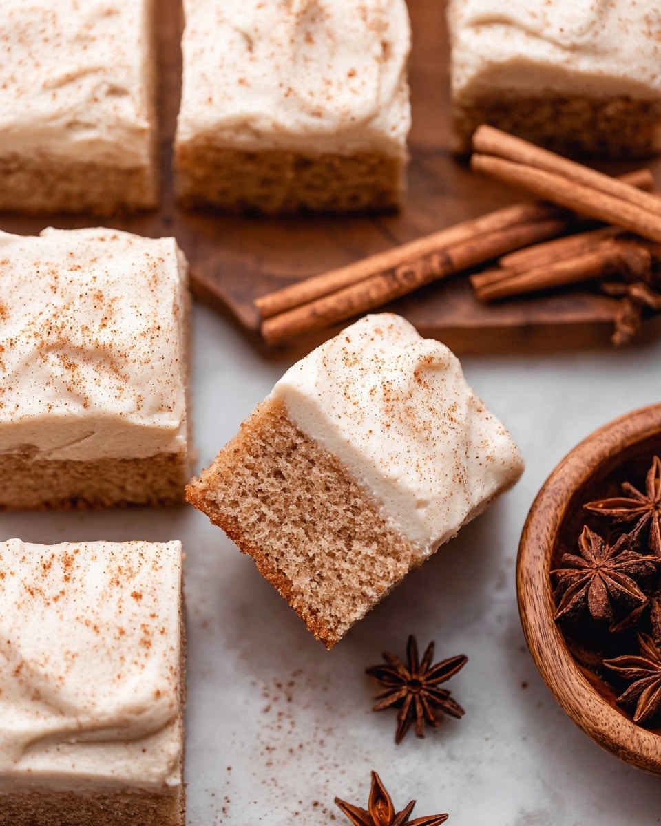 This image shows a group of square cake pieces with two layers each: a light brown cake base with a soft, crumbly texture, and a thick, creamy off-white frosting layer on top sprinkled with fine brown spice powder. One cake square is tilted slightly to show the inside texture, and nearby there is a wooden bowl containing cinnamon sticks and star anise. The entire scene is set on a white marbled surface. photo taken with an iphone --ar 4:5 --v 7