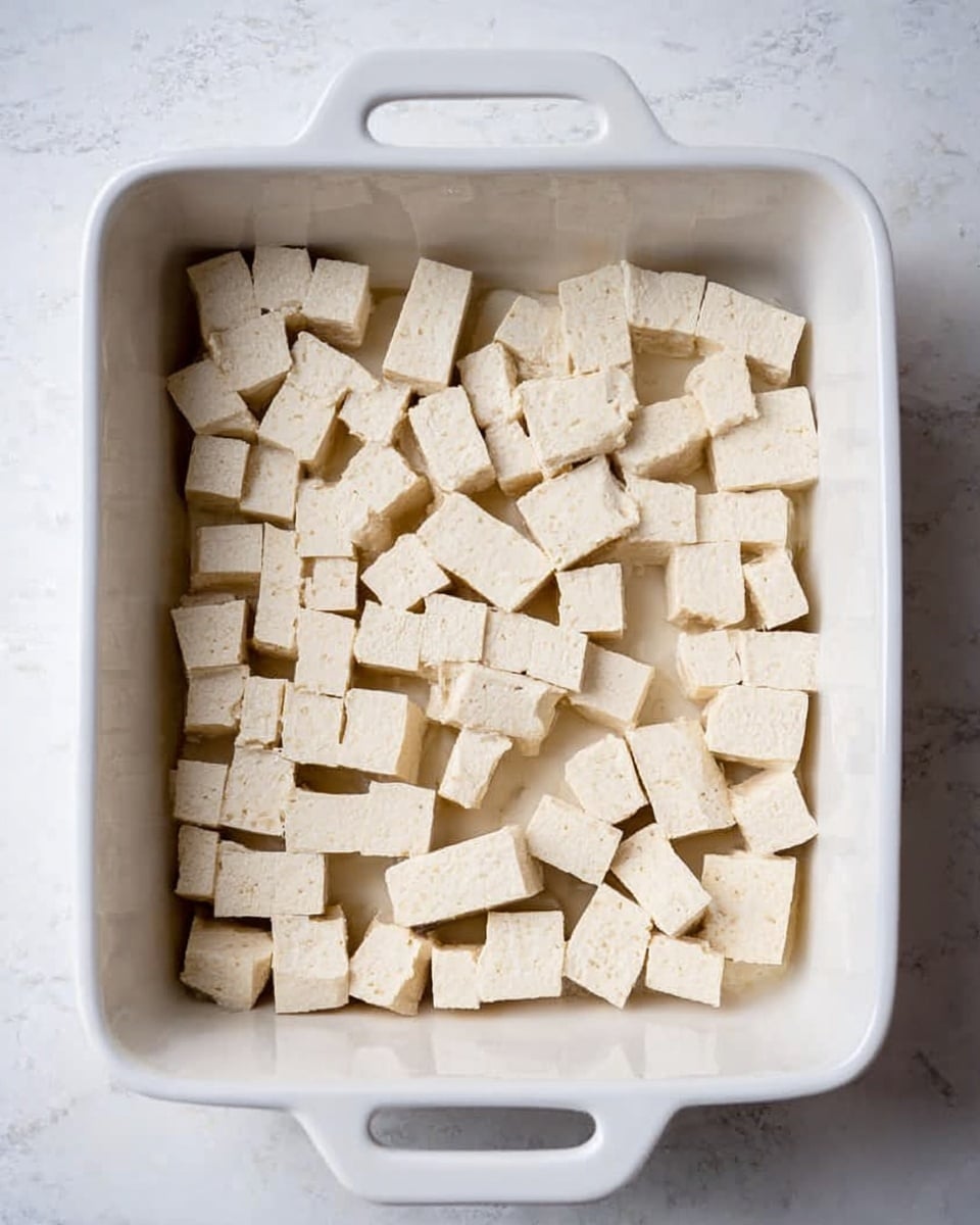 A white baking dish filled with an even layer of light beige tofu cubes. The tofu pieces are arranged neatly, filling the bottom of the dish, with varied sizes but mostly rectangular shapes. The white marbled surface underneath the dish contrasts subtly with the dish’s smooth glossy finish and the tofu’s slightly porous texture. Photo taken with an iphone --ar 4:5 --v 7