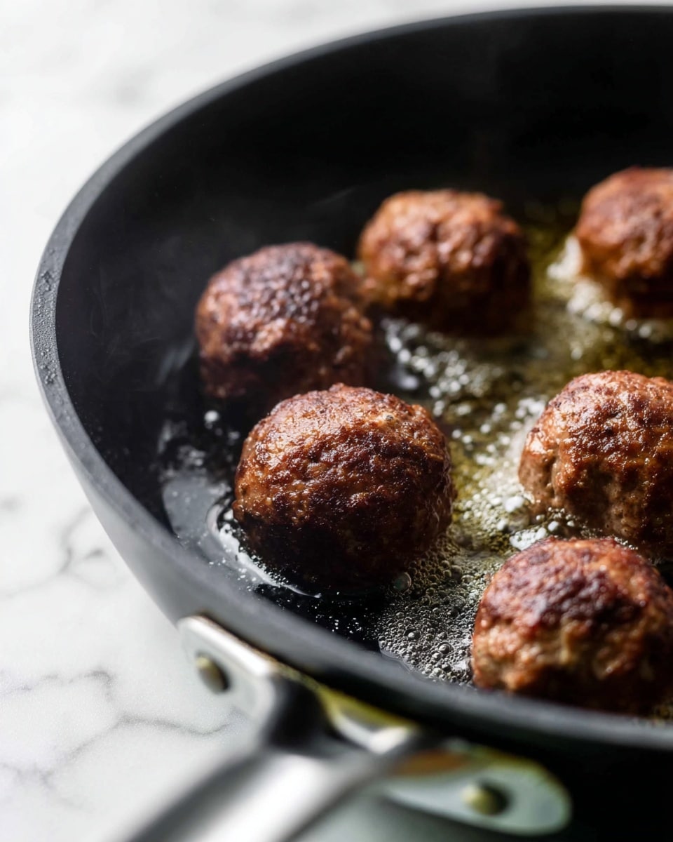 The image shows six round meatballs frying in a black pan with hot oil. Each meatball is brown with a slightly textured surface, showing some parts that are darker and some lighter. The pan's interior has a rough texture with tiny droplets of oil and steam near the meatballs. The pan handle is silver and blurred in the foreground. The background is a white marbled texture. photo taken with an iphone --ar 4:5 --v 7