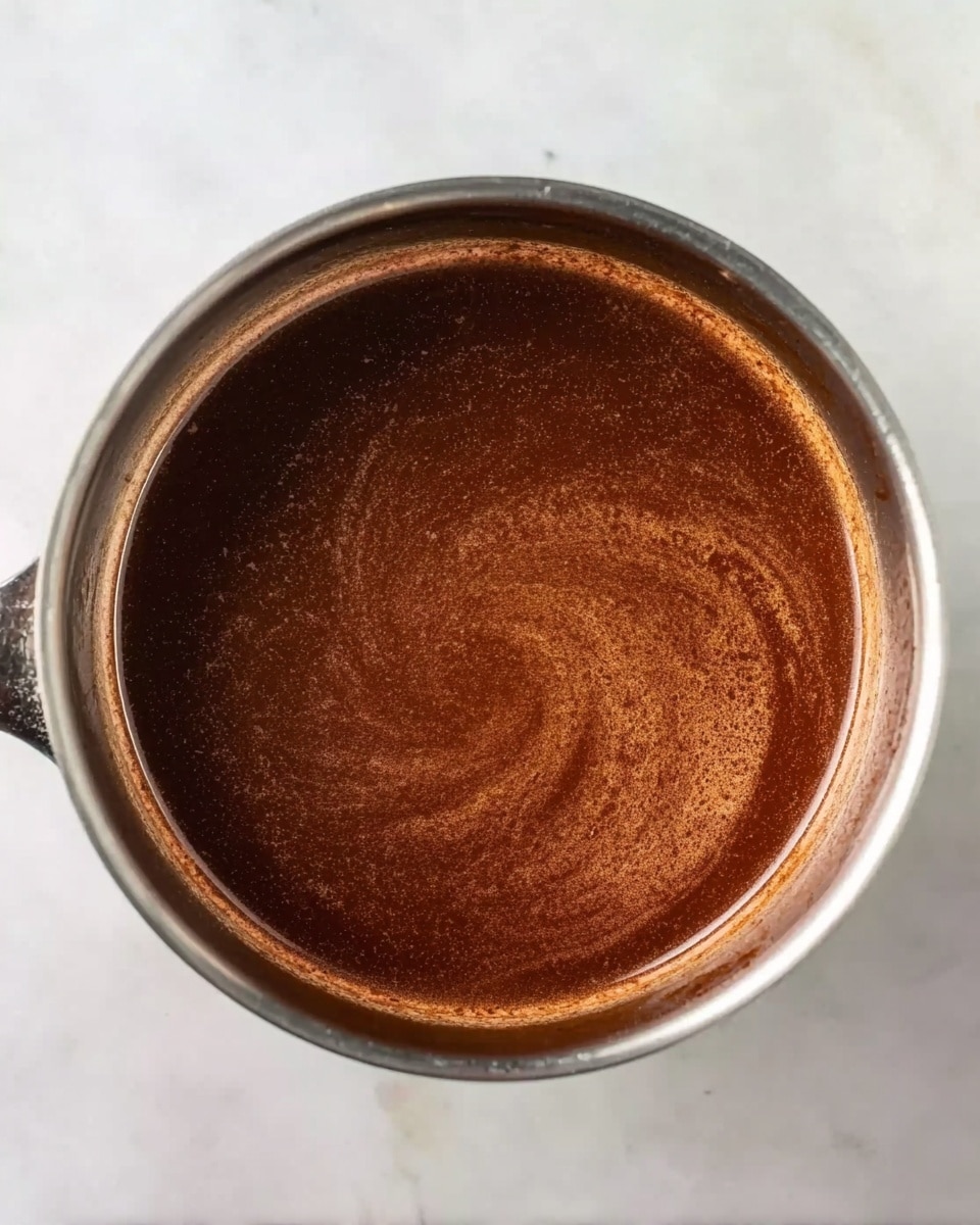 A metal pot filled with a smooth, rich brown sauce that has a slightly shiny and swirled surface inside it. The pot is resting on a white marbled surface, with soft lighting highlighting the texture of the sauce. The sauce layer is uniform without any visible chunks or garnishes, showing a glossy and thick consistency. photo taken with an iphone --ar 4:5 --v 7