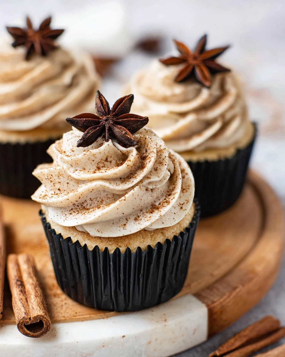 A close-up view of three cupcakes on a wooden round board placed on a white marbled surface. Each cupcake has one layer of light golden cake inside a black liner. On top, there is a thick swirl of light beige frosting sprinkled with a fine dusting of brown cinnamon powder. Each swirl is topped with a single dark brown star anise as decoration. In the background, two cinnamon sticks lie on the white marbled surface. The lighting is bright and soft, making the frosting look creamy and smooth. photo taken with an iphone --ar 4:5 --v 7