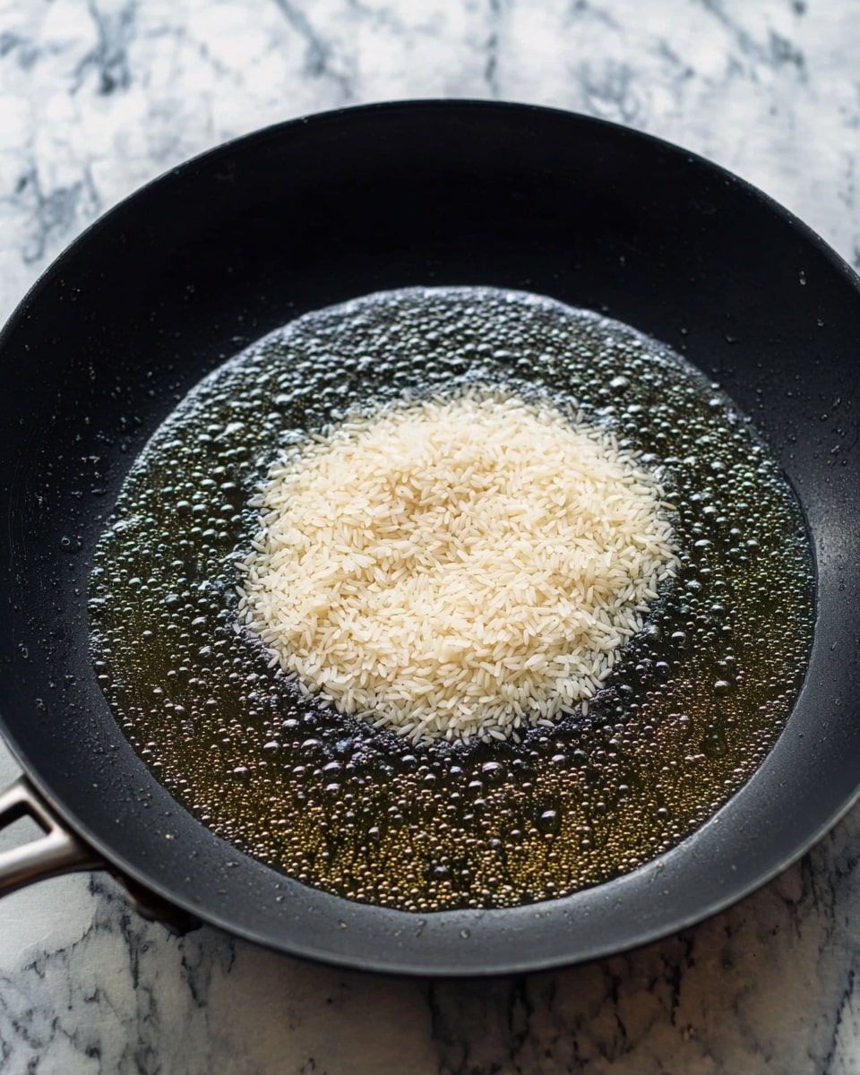 Inside a black frying pan, there is a small pile of uncooked white rice in the center, resting on a layer of melted butter that is spread out around it. The butter is shiny and slightly bubbling, covering the dark surface of the pan. The background shows a white marbled texture beneath the pan photo taken with an iphone --ar 4:5 --v 7