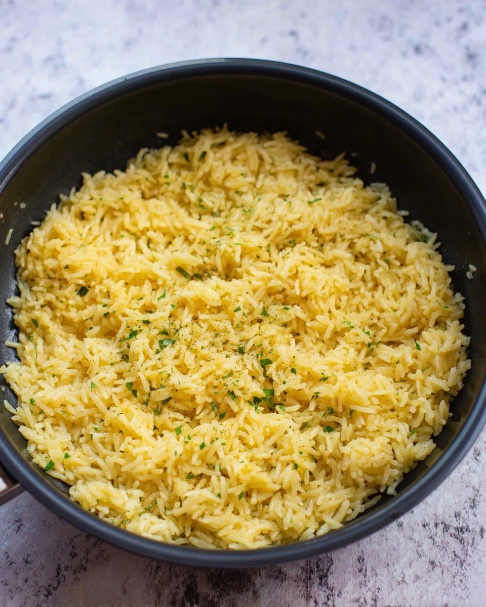 A black pan filled with a thick layer of cooked yellow rice mixed with small green herb bits, the rice looks fluffy and slightly oily with a soft texture. The pan is placed on a white marbled surface. Photo taken with an iphone --ar 4:5 --v 7