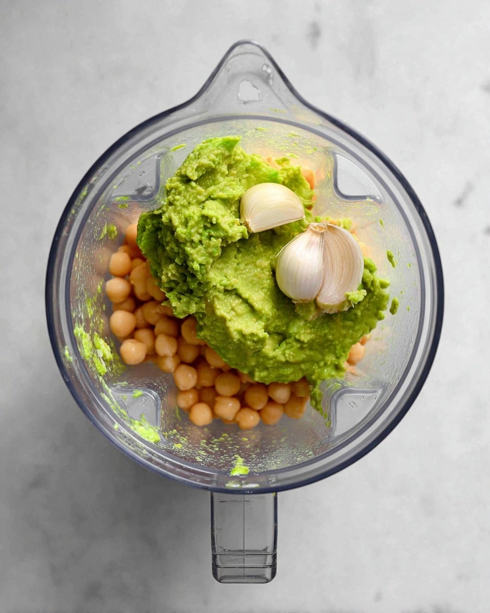 A clear blender jar is shown from above on a white marbled surface, filled with three main layers of ingredients. The bottom layer consists of light beige chickpeas. On top of that, there is a generous dollop of green mashed avocado with a creamy texture. Sitting on the avocado is one whole clove of white garlic. The blender is transparent, and the scene is well-lit to show the fresh colors of the ingredients clearly. photo taken with an iphone --ar 4:5 --v 7