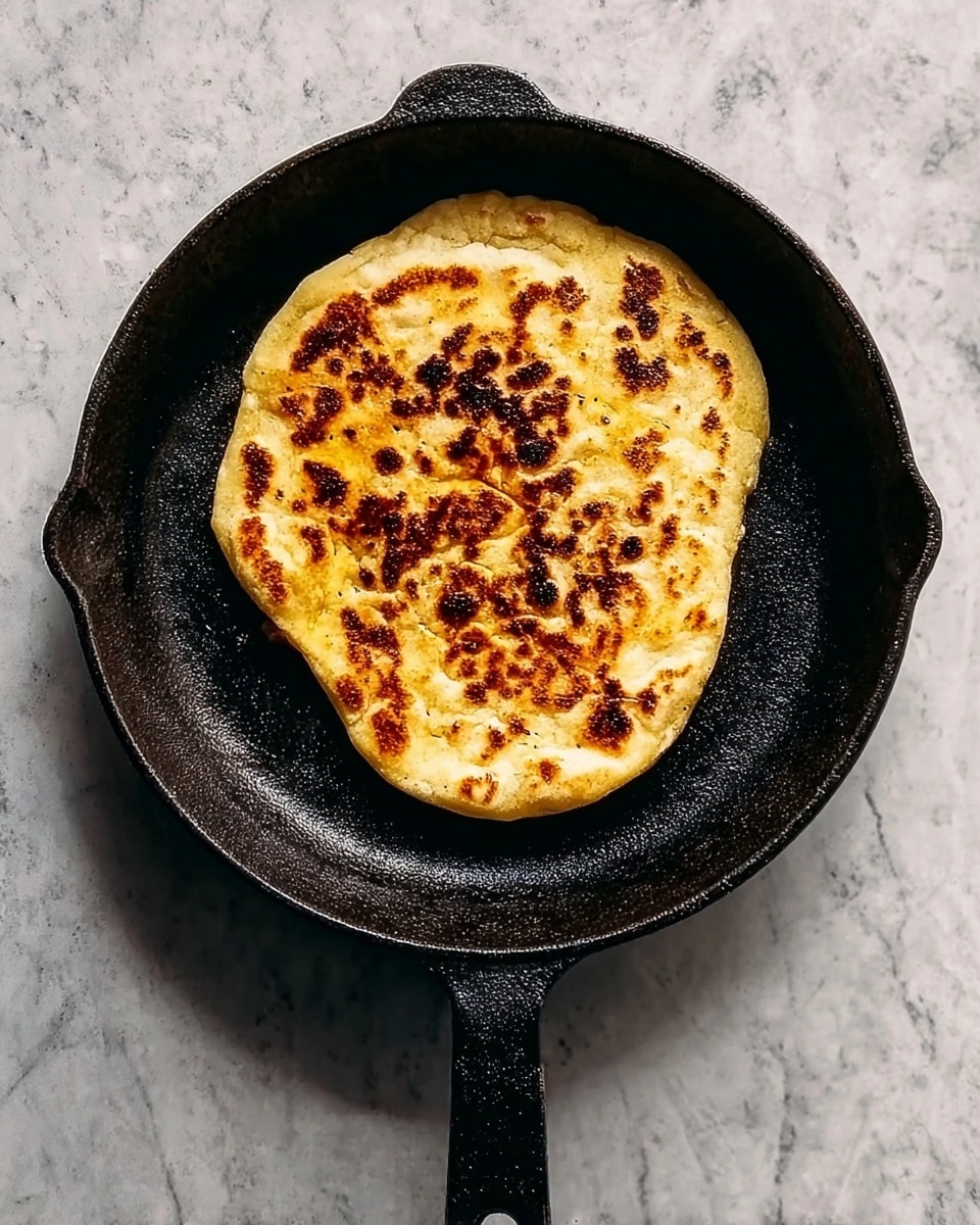 A single flatbread with an uneven round shape lies in the center of a black cast iron pan. The flatbread has a golden-yellow color with darker brown char spots scattered mostly in the middle area, showing a crispy, slightly rough texture. The pan has a long handle with a hole at the end, and the background is a white marbled texture. The flatbread appears thick and soft with a slightly uneven surface. Photo taken with an iphone --ar 4:5 --v 7