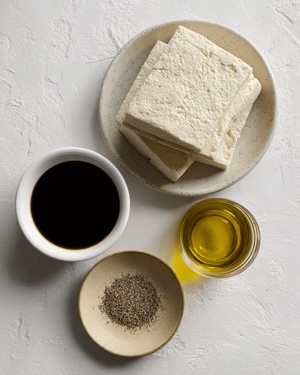 The image shows a block of pale, slightly textured tofu resting in a shallow speckled white bowl at the top right. Below and slightly left, a small white bowl holds a deep, dark liquid, likely soy sauce, with a smooth surface reflecting light. Next to it on the right is a small clear glass container filled with golden olive oil. At the bottom center, there is a small round beige plate with coarse black pepper sprinkled on it. All items sit on a white marbled surface. Photo taken with an iphone --ar 4:5 --v 7
