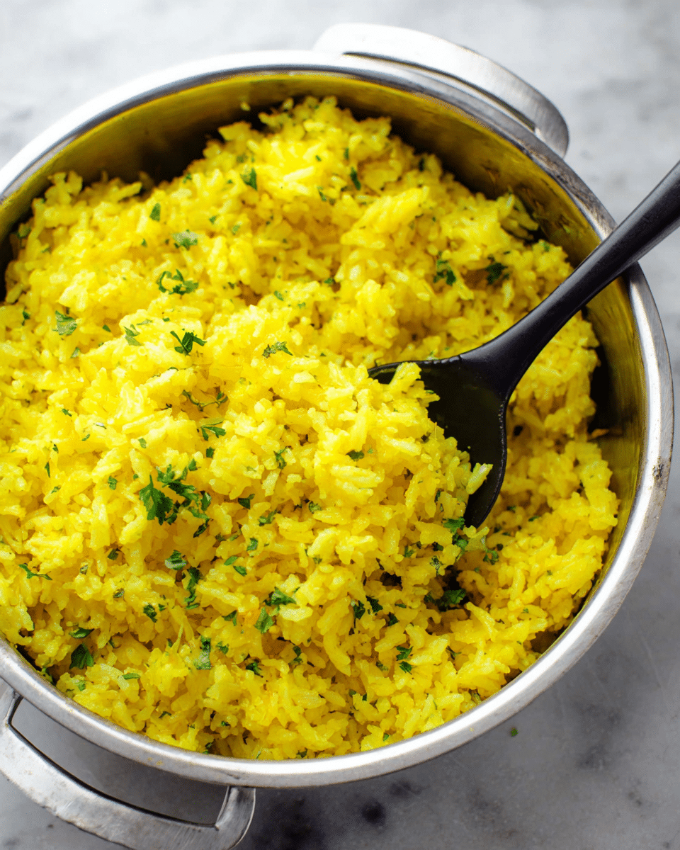 The image shows a close-up of a shiny silver pot filled with bright yellow rice. The rice has a fluffy texture and is mixed with small bits of green herbs scattered on top, adding color contrast. There is a black spoon resting inside the pot, partly buried in the rice, showing the grains clearly. The pot is placed on a white marbled surface, giving a clean and fresh look. photo taken with an iphone --ar 4:5 --v 7