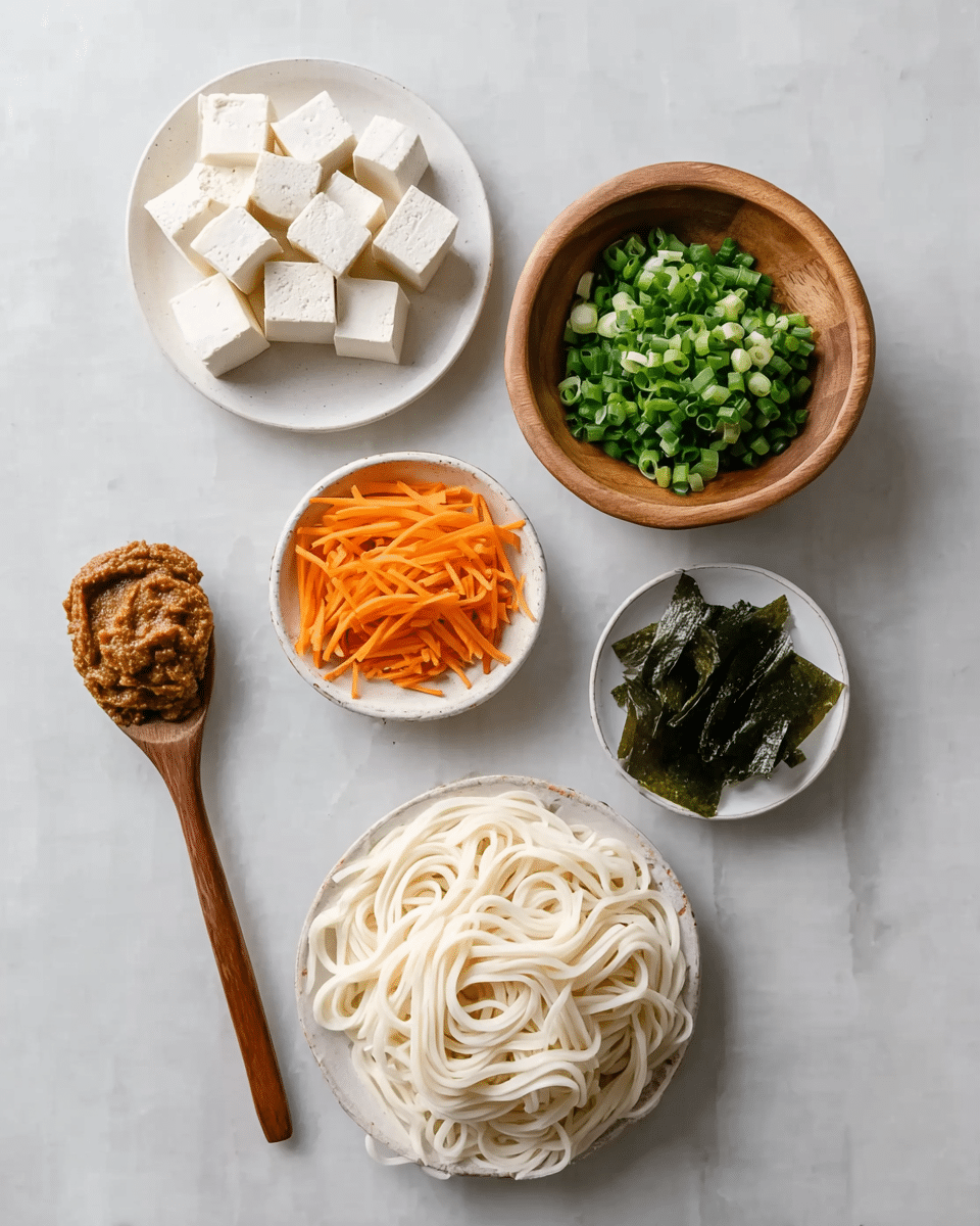 The image shows six groups of food on a white marbled surface. In the top left is a white plate with small white cubes of tofu. To the right is a wooden bowl filled with chopped green onions. Below is a white bowl with thin orange carrot strips. Next to it on the right is a small white bowl holding thin strips of dark green seaweed. Below is a white plate with thick white udon noodles in a loose pile. At the bottom left, a wooden spoon holds a thick brown paste. The arrangement is neat and the colors are bright and fresh. photo taken with an iphone --ar 4:5 --v 7