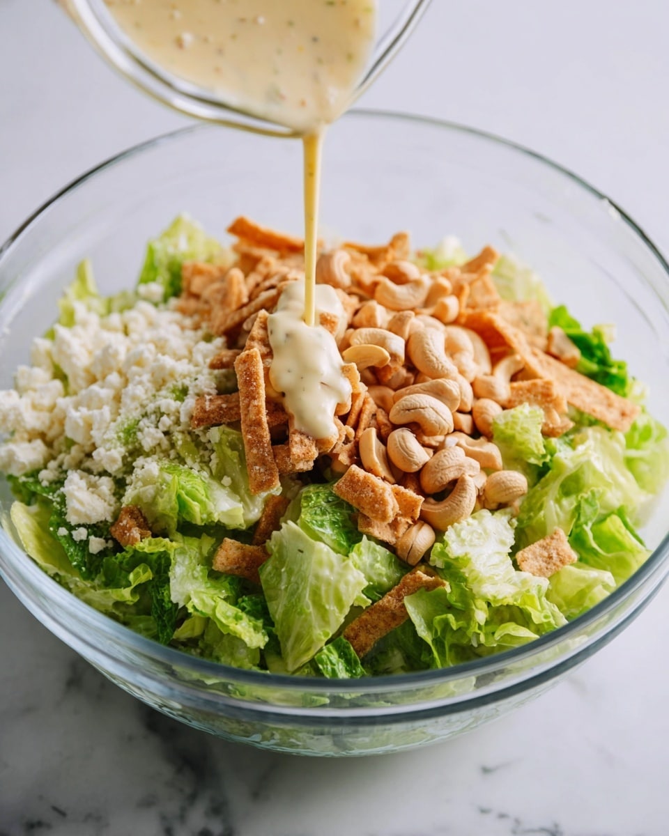 A clear glass bowl contains a layered salad on a white marbled surface. The bottom layer is fresh, bright green lettuce leaves, roughly torn. On top of the lettuce, there is a pile of crumbled white cheese on one side, crunchy tan-colored fried tortilla strips on another side, and light brown cashew nuts scattered among the lettuce. Creamy, pale yellow salad dressing is being poured over the top from above, covering parts of the cheese, tortilla strips, and lettuce. The image shows vibrant colors and texture contrasts, with the glass bowl allowing full view of the ingredients inside. Photo taken with an iphone --ar 4:5 --v 7