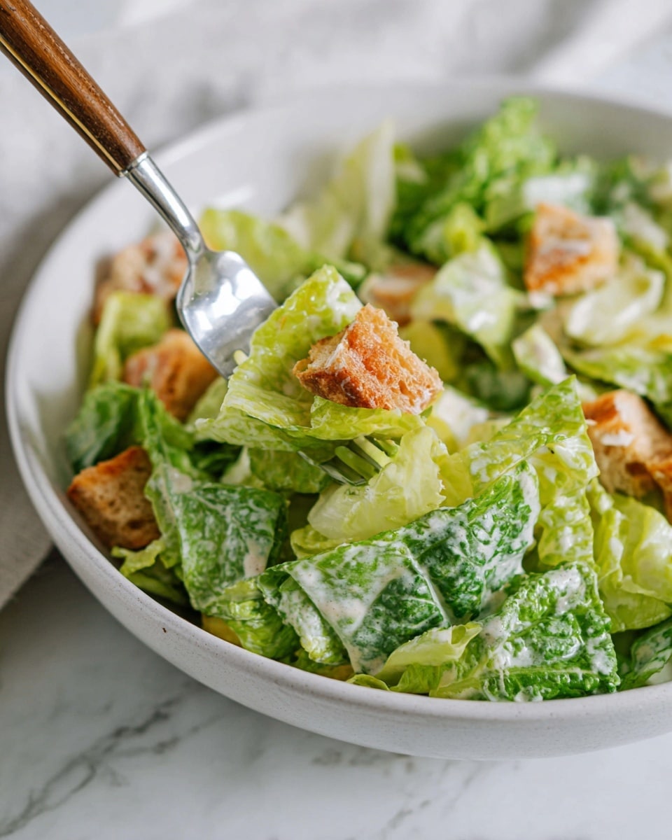 A white bowl filled with a fresh Caesar salad showing three main layers: the bottom layer has bright green romaine lettuce leaves with a crisp texture, the middle layer contains light brown croutons with a crunchy look, and the top layer is coated with a creamy white dressing that adds a smooth shine across the ingredients. A silver fork with a wooden handle lifts a bite that combines all these layers, with pieces of lettuce, dressing, and croutons clearly visible. The bowl sits on a white marbled surface that gives a clean and fresh background to the dish, photo taken with an iphone --ar 4:5 --v 7