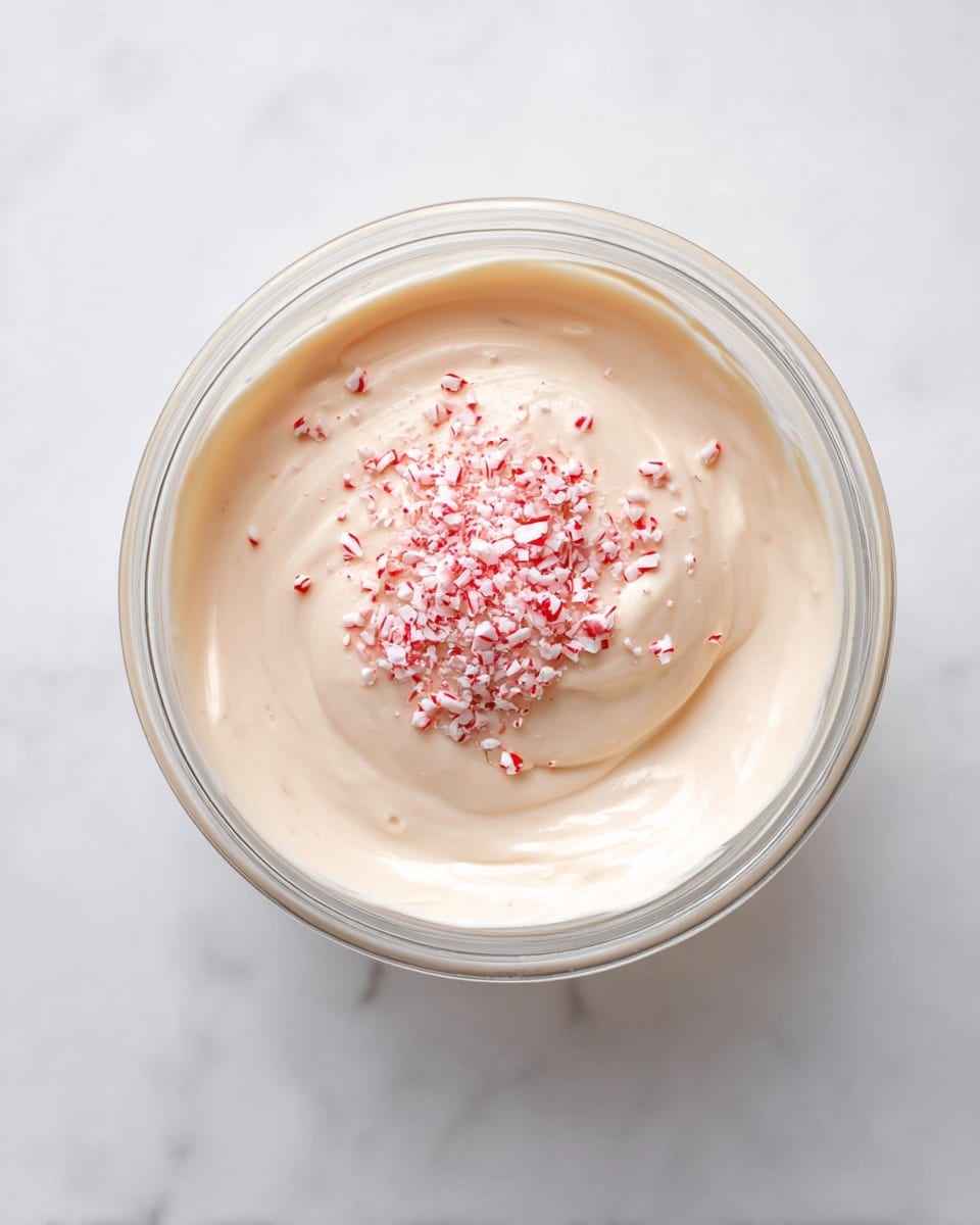 A clear glass bowl sits on a white marbled surface, filled with one smooth, creamy layer of light beige mixture. On top of this creamy layer is a small pile of finely crushed red and white peppermint candy bits, adding a textured contrast at the center of the bowl. The mixture looks soft and thick, with visible gentle swirls showing slight movement. photo taken with an iphone --ar 4:5 --v 7