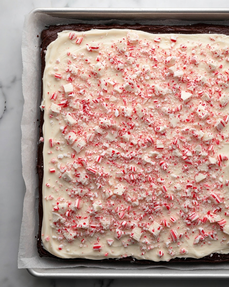 A rectangular tray lined with parchment paper holds a layered treat. The bottom layer is dark chocolate spread smoothly, covering the entire surface except the edges. On top, a thick and creamy white layer is evenly spread, almost reaching the edges of the tray. The top is covered with crushed peppermint candy pieces in red and white, scattered densely and unevenly, giving a crunchy texture look. Some larger peppermint chunks stand out among the finer crushed bits. The tray is on a white marbled surface. photo taken with an iphone --ar 4:5 --v 7