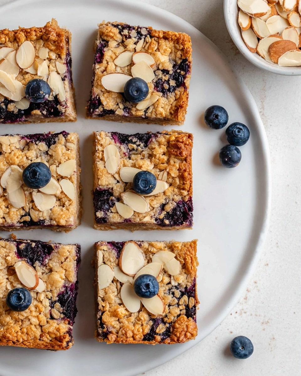 The image shows six square oat bars arranged in two rows of three on a white plate. Each bar has a golden-brown oatmeal base with dark purple blueberries scattered inside and on top. The top layer is sprinkled with light beige, thin almond slices. Two fresh blueberries sit on the white marbled surface near the plate. In the top right, part of a white bowl with extra almond slices is visible. The oat bars have a slightly rough, crumbly texture, and the edges are firm. photo taken with an iphone --ar 4:5 --v 7