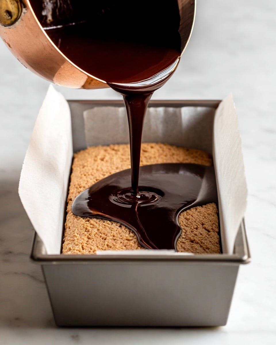 A close-up shows a shiny dark chocolate liquid being poured from a copper saucepan onto a thick, light brown textured layer inside a metal loaf pan lined with parchment paper. The light brown layer fills the bottom of the pan and looks rough and uneven. The shiny chocolate layer spreads on top, creating a smooth, glossy dark contrast over the rough base. The pan sits on a white marbled surface in soft light. photo taken with an iphone --ar 4:5 --v 7