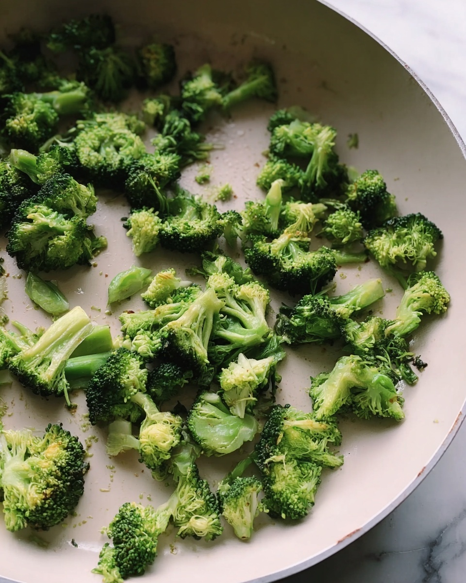 The image shows a close-up of small green broccoli pieces scattered inside a white frying pan. The broccoli pieces have a mix of dark green tree-like tops and lighter green stalks with a slightly soft texture. The pan's smooth white surface contrasts with the rough texture of the broccoli. The background shows a blurred white marbled texture. photo taken with an iphone --ar 4:5 --v 7