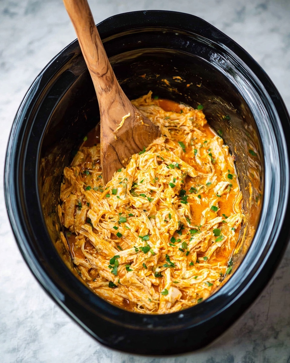Slow Cooker Buffalo Chicken Sweet Potatoes Recipe 4 Inside a black slow cooker, there is one layer of shredded chicken mixed with an orange sauce, making the texture look soft and creamy. Small green chopped herbs are scattered evenly across the chicken, adding bits of color contrast. A wooden spoon rests inside, with the spoon’s bowl slightly buried in the chicken, showing the mix's thickness and smoothness. The slow cooker is set on a white marbled surface. photo taken with an iphone --ar 4:5 --v 7