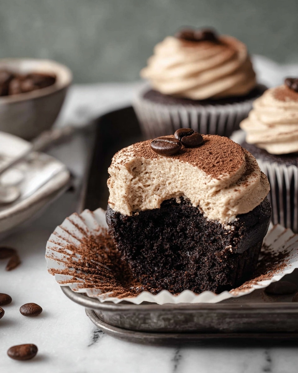 The image shows a close-up of a dark chocolate cupcake with a creamy light brown frosting layer on top, dusted with cocoa powder and topped with two coffee beans. The cupcake liner is white with ridges, and the cupcake itself has a moist, crumbly texture visible where a bite has been taken. The cupcake is placed on a dark metal tray, with a white marbled texture in the background, and there are coffee beans scattered around. In the background, two more cupcakes with the same frosting and decoration are visible. photo taken with an iphone --ar 4:5 --v 7