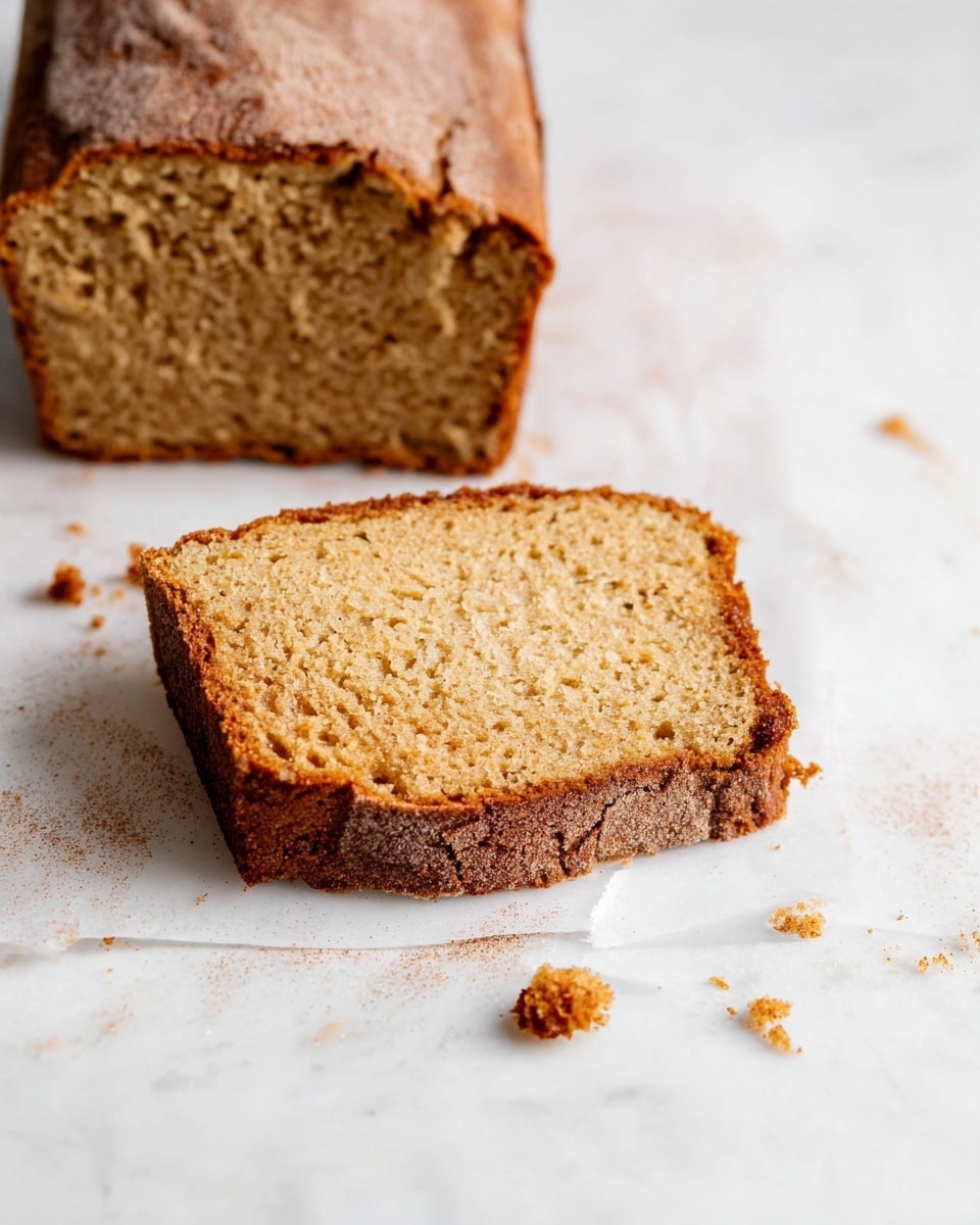 A slice of brown loaf cake with a slightly crumbly texture rests on a white marbled surface, alongside a larger piece of the same loaf. The cake has a light golden brown color with a darker, slightly cracked top dusted with a light layer of cinnamon or sugar. The slice shows a dense but soft interior with small air holes throughout. Some crumbs are scattered around the loaf pieces on the white marbled surface. photo taken with an iphone --ar 4:5 --v 7