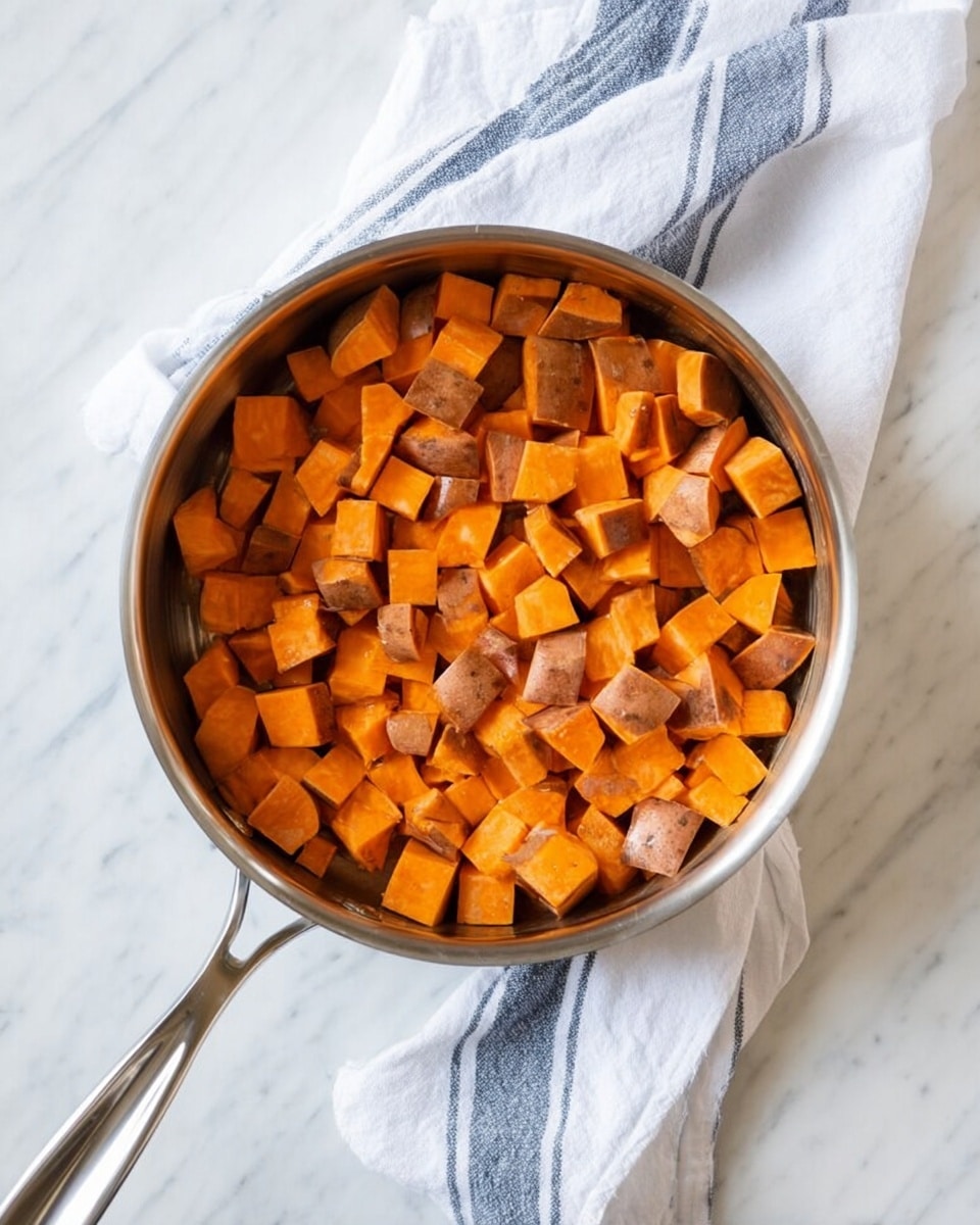 A silver saucepan is filled with cubes of sweet potatoes in orange and brown skin colors, fully covering the pan. The pan has a long handle and is placed on a white marbled surface. To the right of the pan, there is a white cloth with blue stripes loosely folded. The image has soft natural light and a simple clean look. photo taken with an iphone --ar 4:5 --v 7