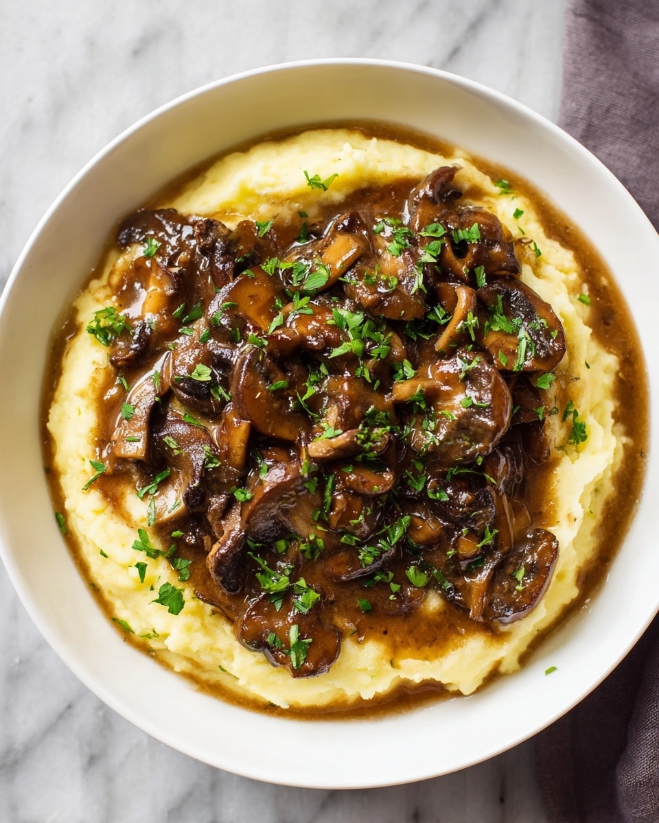 A white bowl holds a dish with two main layers; the bottom layer is creamy mashed potatoes in a pale yellow color with a soft, smooth texture, filling the bowl evenly. On top, a thick layer of dark brown mushrooms in a rich sauce covers the center, the mushrooms having a glossy, slightly chunky texture and irregular shapes. Fresh green chopped herbs are sprinkled over the mushrooms, adding a pop of color. The bowl sits on a white marbled surface. Photo taken with an iphone --ar 4:5 --v 7