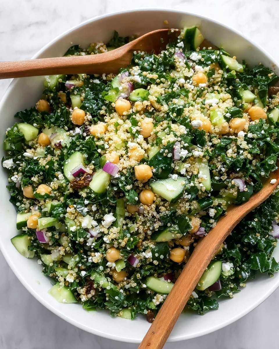 The image shows a white bowl filled with a mixed salad. The salad has three main layers: the bottom layer is full of chopped dark green kale leaves, the middle layer has small pale yellow quinoa grains evenly spread out, and scattered throughout are light beige chickpeas and golden raisins. On top, there are small pieces of chopped red onion, green cucumber chunks, and tiny white crumbles of cheese. Two wooden spoons rest inside the bowl, partly covered by the salad. The bowl is placed on a white marbled surface photo taken with an iphone --ar 4:5 --v 7
