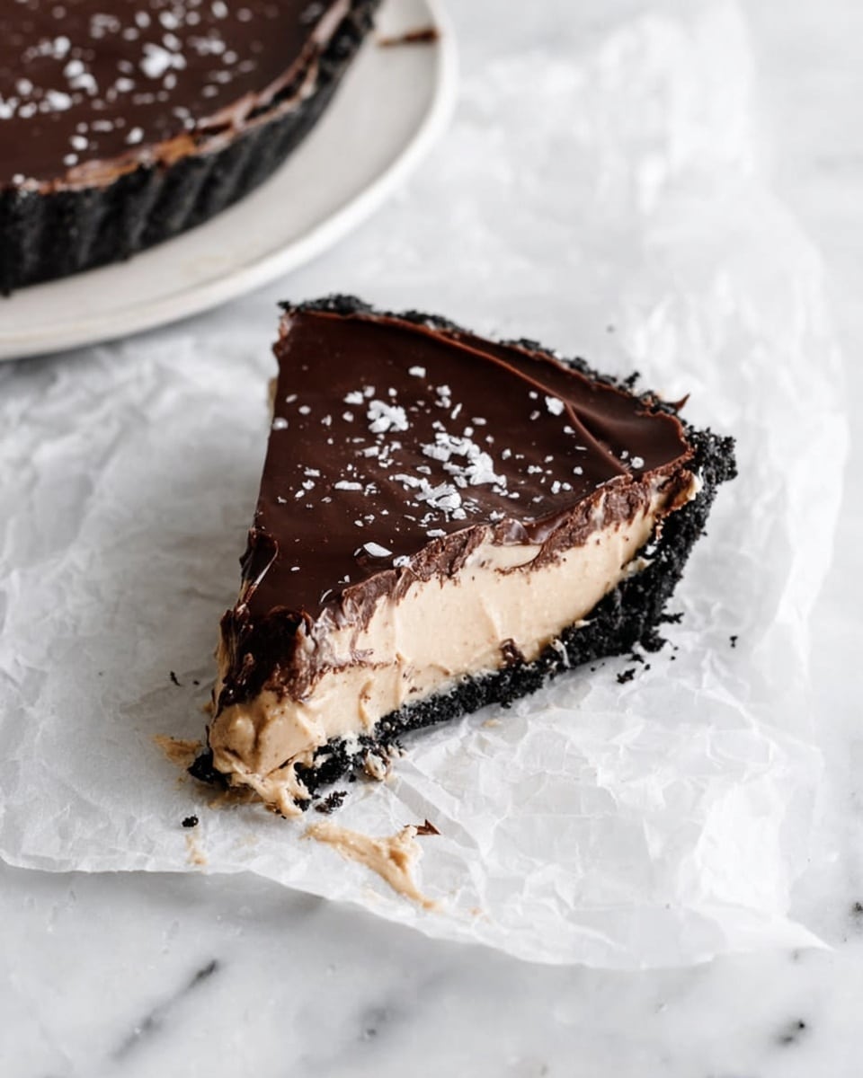 A single slice of pie with three visible layers sits on a piece of crumpled white paper, placed on a white marbled surface. The bottom layer is a thick, crumbly dark crust, almost black in color. Above it is a lighter beige layer with a smooth and creamy texture, spilling slightly onto the paper. The top layer is a glossy dark chocolate, smooth with a few cracks, sprinkled lightly with white flakes. In the upper left corner of the image, part of the remaining pie on a white plate is visible on the same white marbled surface. Photo taken with an iphone --ar 4:5 --v 7
