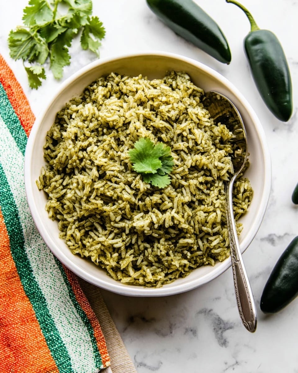 A white bowl filled with one layer of green rice that has a slightly grainy texture, garnished with a few small cilantro leaves in the center. A silver spoon rests inside the rice on the right side. The bowl sits on a white marbled surface with a colorful green, orange, and white striped cloth to the left and two dark green poblano peppers on the top right. There is also a small sprig of cilantro near the top left. photo taken with an iphone --ar 4:5 --v 7