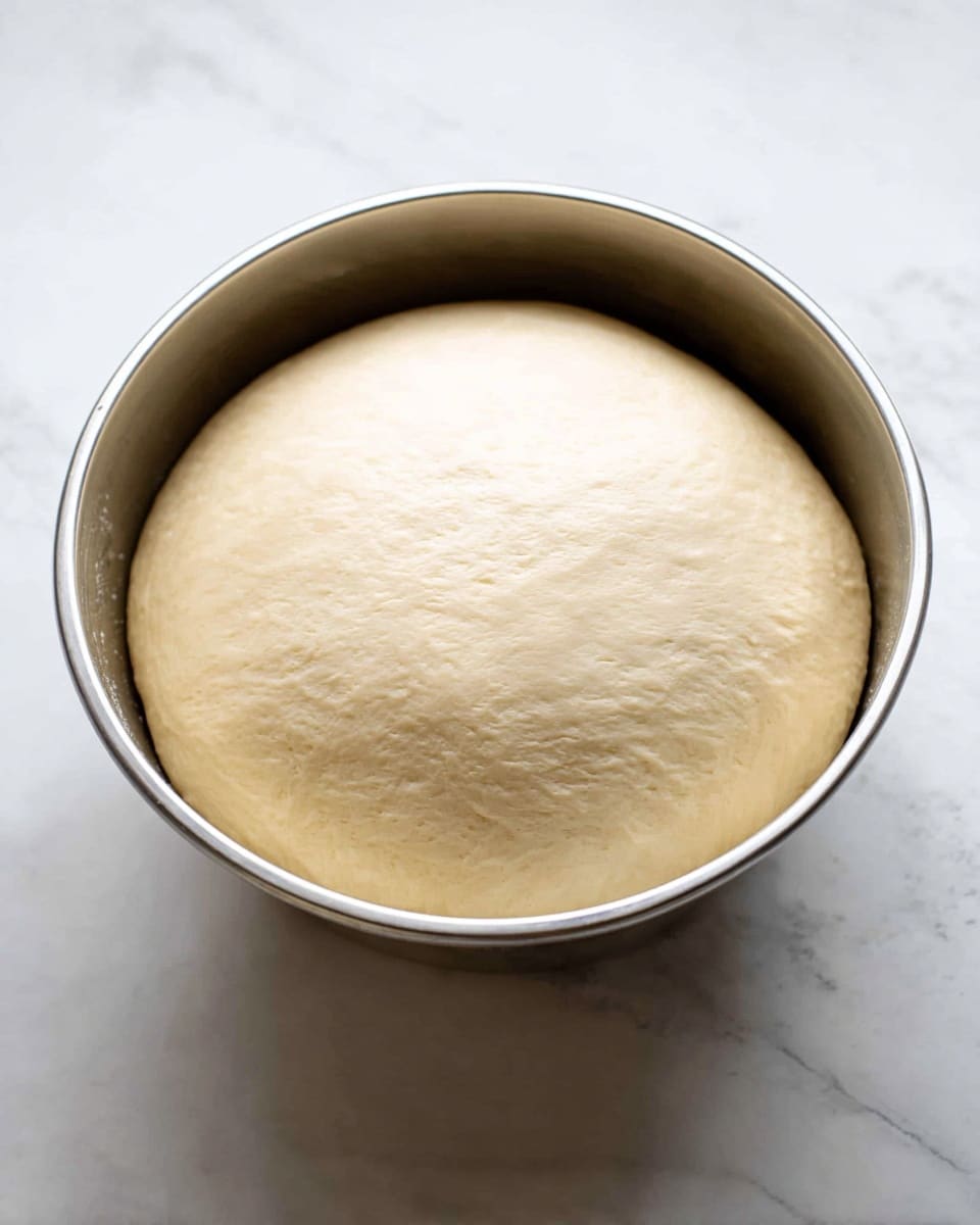 A silver bowl filled with one large round dough ball that has grown and risen, showing a smooth, light beige surface with a soft texture. The bowl is placed on a white marbled surface, and the dough fills nearly the whole inside space, creating a slight dome shape. The light shines softly on the dough, highlighting its smoothness. photo taken with an iphone --ar 4:5 --v 7