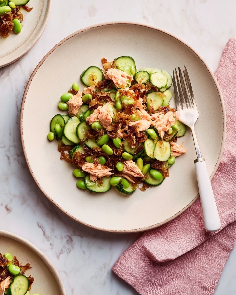 The dish is served on a white plate with a slightly raised edge, filled with a mix of several layers. The base layer consists of small, round slices of fresh cucumber, bright green in color with a smooth texture. Scattered among the cucumber slices are glossy, green edamame beans. On top of these, there are chunks of light pink salmon, flaky and tender. Over the salmon and vegetables, there is a layer of finely chopped caramelized onions or similar browned ingredients, adding a dark brown, slightly crispy texture. The plate is placed on a white marbled surface, accompanied by a white-handled fork resting on the edge of the plate, and a soft pink cloth napkin to the right side. Photo taken with an iphone --ar 4:5 --v 7